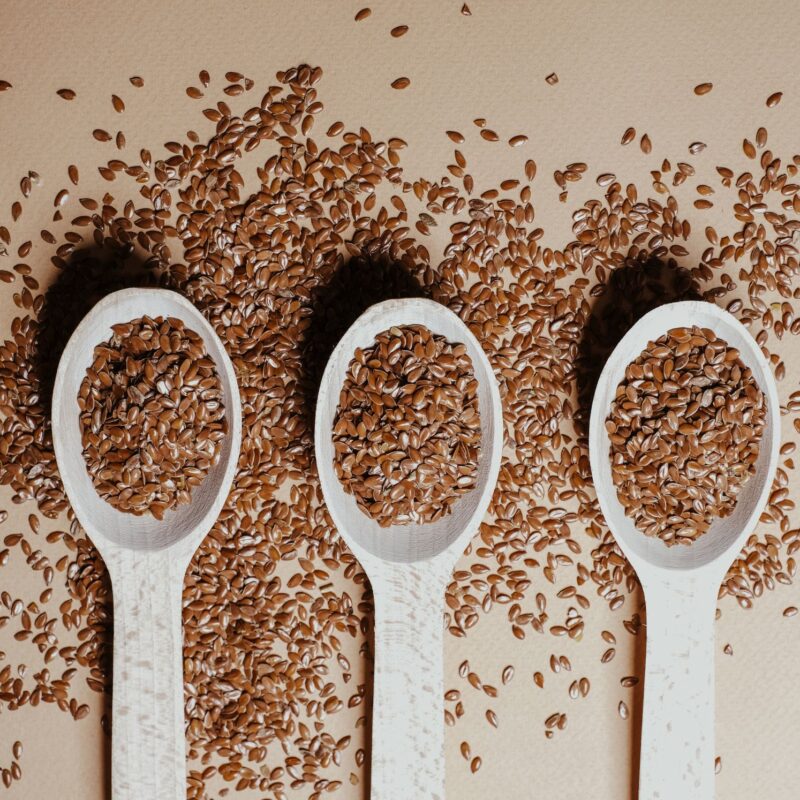Three wooden spoons filled with flaxseed, a rich source of omega 3, are placed side by side on a beige surface, with additional flaxseeds scattered around them.