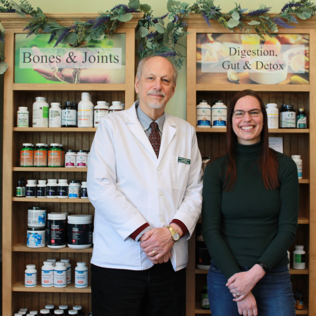 A man in a white lab coat and a woman in a green sweater stand smiling in front of shelves with vitamin bottles. Signs above read “Bones & Joints” and “Digestion, Gut & Detox.” Greenery decorates the shelves.