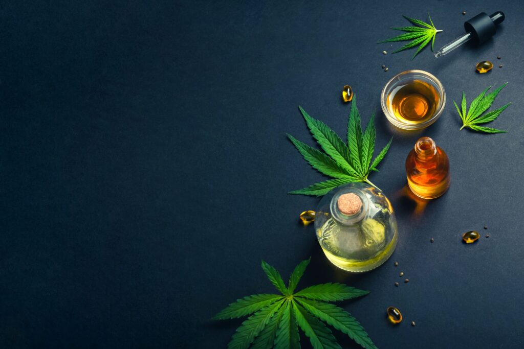 Bottles of full spectrum CBD oil, capsules, a dropper, and cannabis leaves are arranged on a dark surface with some scattered seeds and leaves.