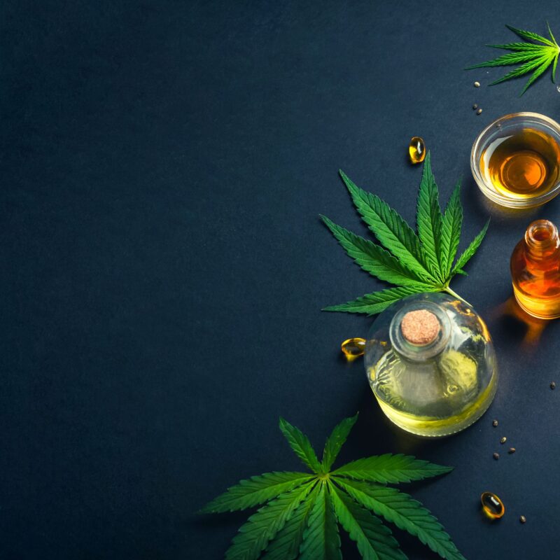 Bottles of full spectrum CBD oil, capsules, a dropper, and cannabis leaves are arranged on a dark surface with some scattered seeds and leaves.