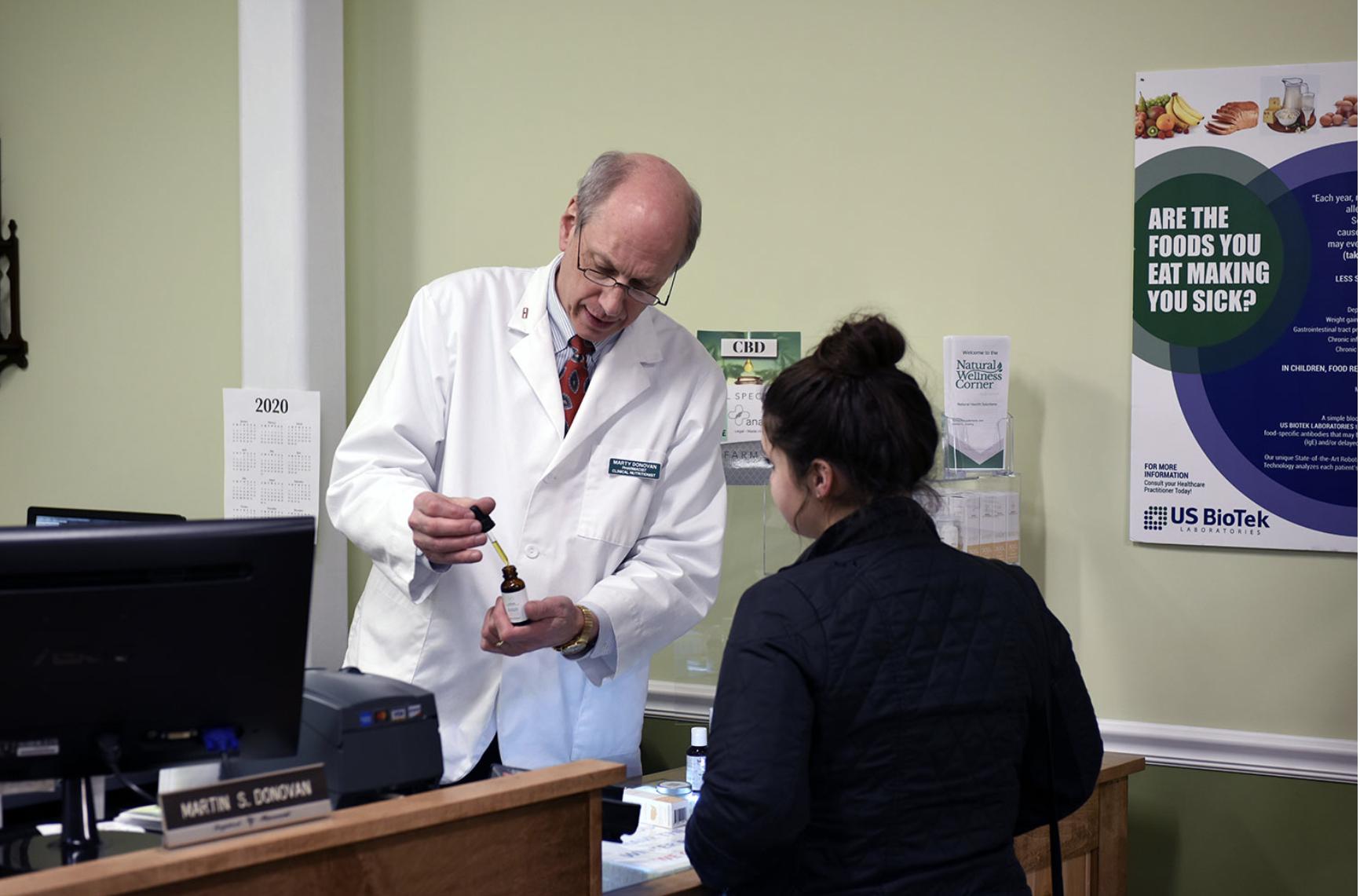 A pharmacist in a white coat explains medication to a customer at a pharmacy counter. The customer listens attentively. A computer, pamphlets, and a health-related poster are visible in the background.
