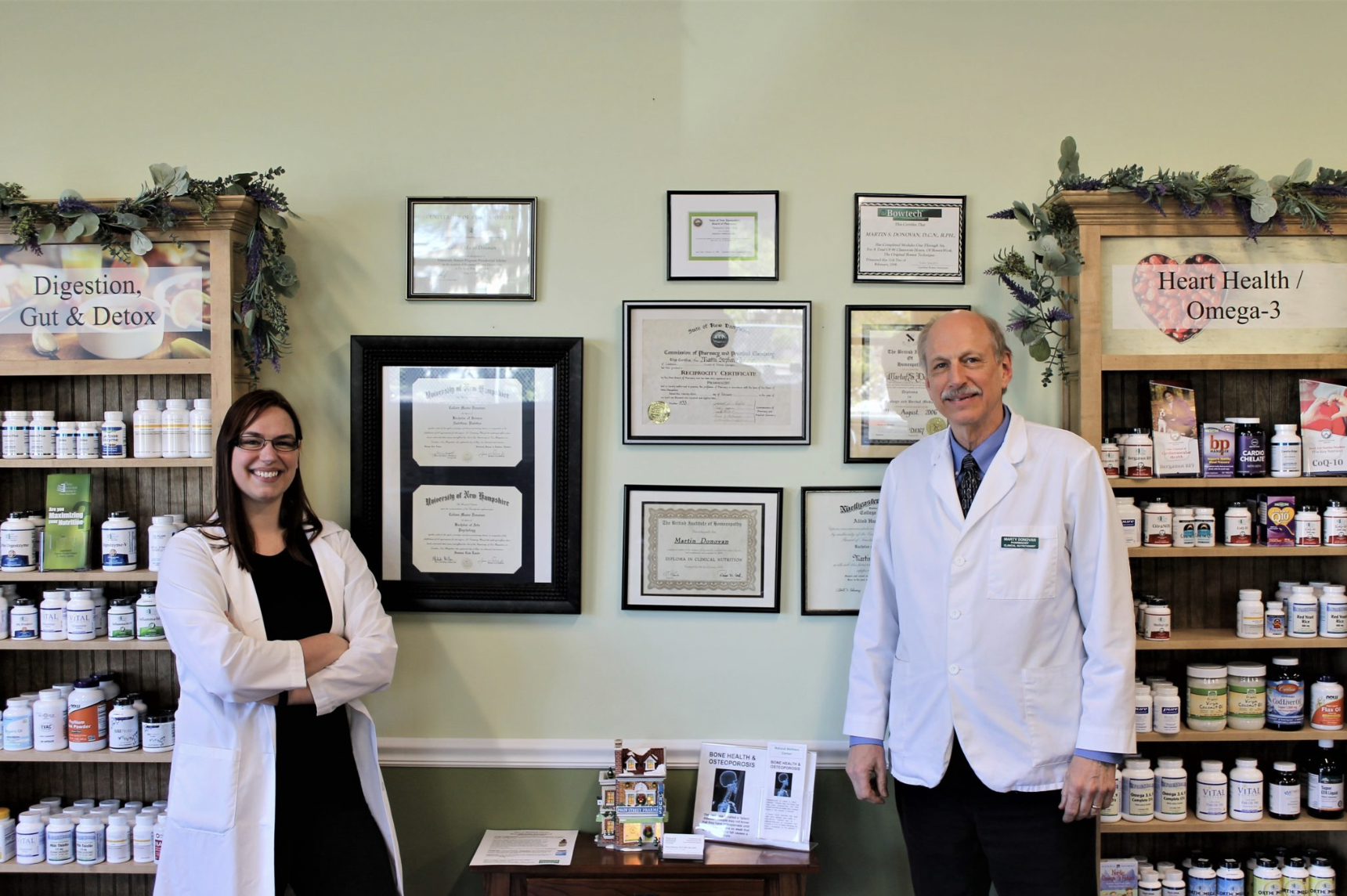 Two people in white lab coats stand in a supplement store, surrounded by shelves of vitamin bottles. Framed certificates hang on the wall behind them, and signs about digestion and heart health are visible on the shelves.