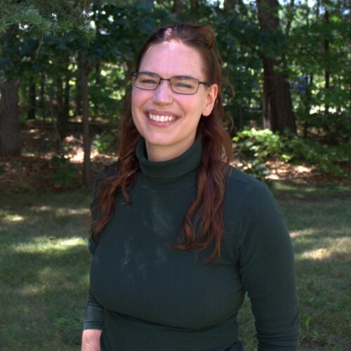 A picture of Colleen Donovan, CNS, a woman with long brown hair and glasses smiles outdoors, wearing a dark green ribbed turtleneck. She stands in a sunlit wooded area with green trees and grass in the background, embodying the approachable professionalism of a nutritionist in Concord, NH.